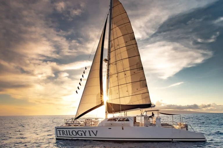 A sailboat named "Trilogy IV" sailing on the ocean at sunset with large sails and a partly cloudy sky.