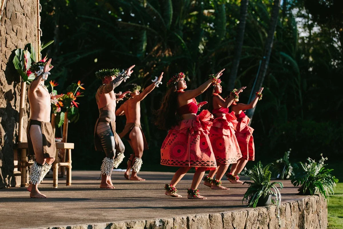 Hula dancers performing on an outdoor stage in traditional Hawaiian attire, surrounded by lush greenery and tropical plants.
