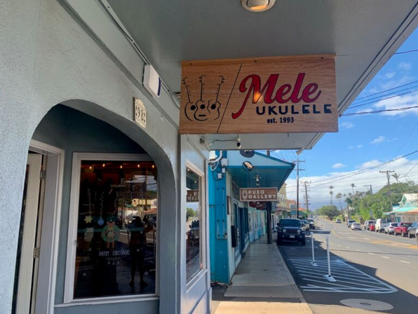 Street view with storefronts, including Mele Ukulele store with a wooden sign, on a sunny day with a clear blue sky, parked cars, and a pedestrian sidewalk.