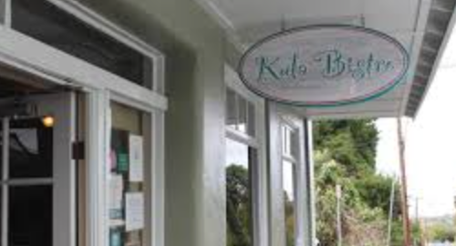 A storefront with a sign that reads 'Kate Baker,' with large glass windows and a door, and trees visible in the background.
