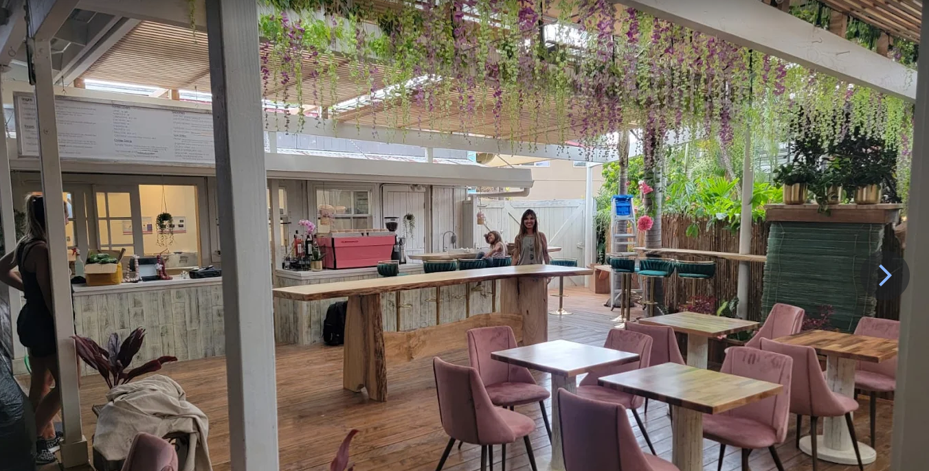 A cozy outdoor cafe with pink chairs, wooden tables, hanging purple flowers, and lush green plants, with two women standing and chatting.