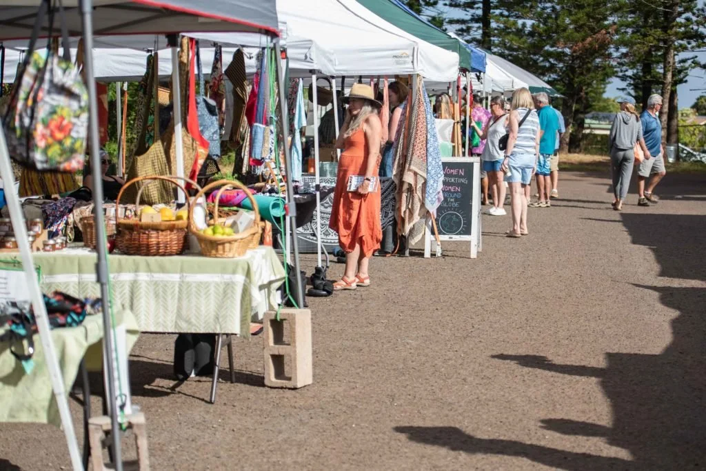 People browsing outdoor market stalls under white tents with various craft and food items, on a dirt pathway on a sunny day.