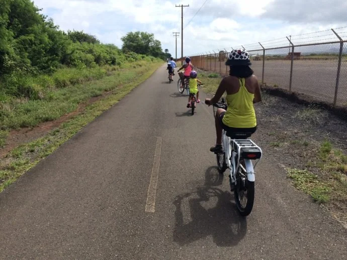 Group of people, including children, riding bicycles on a paved path next to a chain-link fence and greenery on a cloudy day.