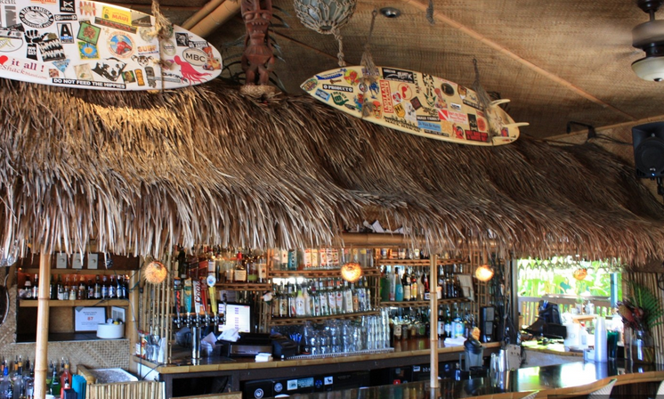 Bar area of a tropical-themed restaurant or bar with thatched roof, surfboards decorated with stickers hanging from the ceiling, and shelves stocked with bottles of alcohol.