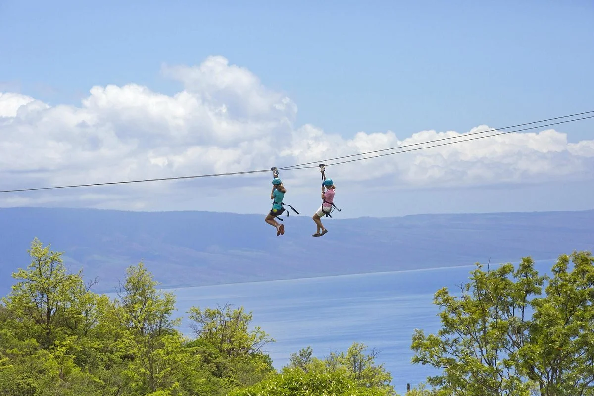 Two children wearing helmets and harnesses are zip lining over a body of water with green trees in the foreground and mountains and clouds in the background.