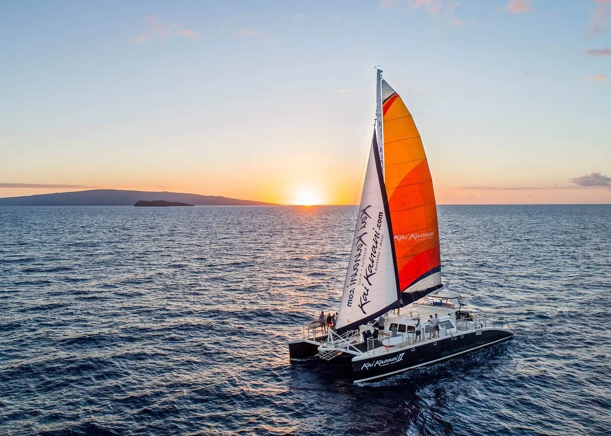 A sailboat with orange, yellow, and white sails on the ocean during sunset, with a landmass in the distance.
