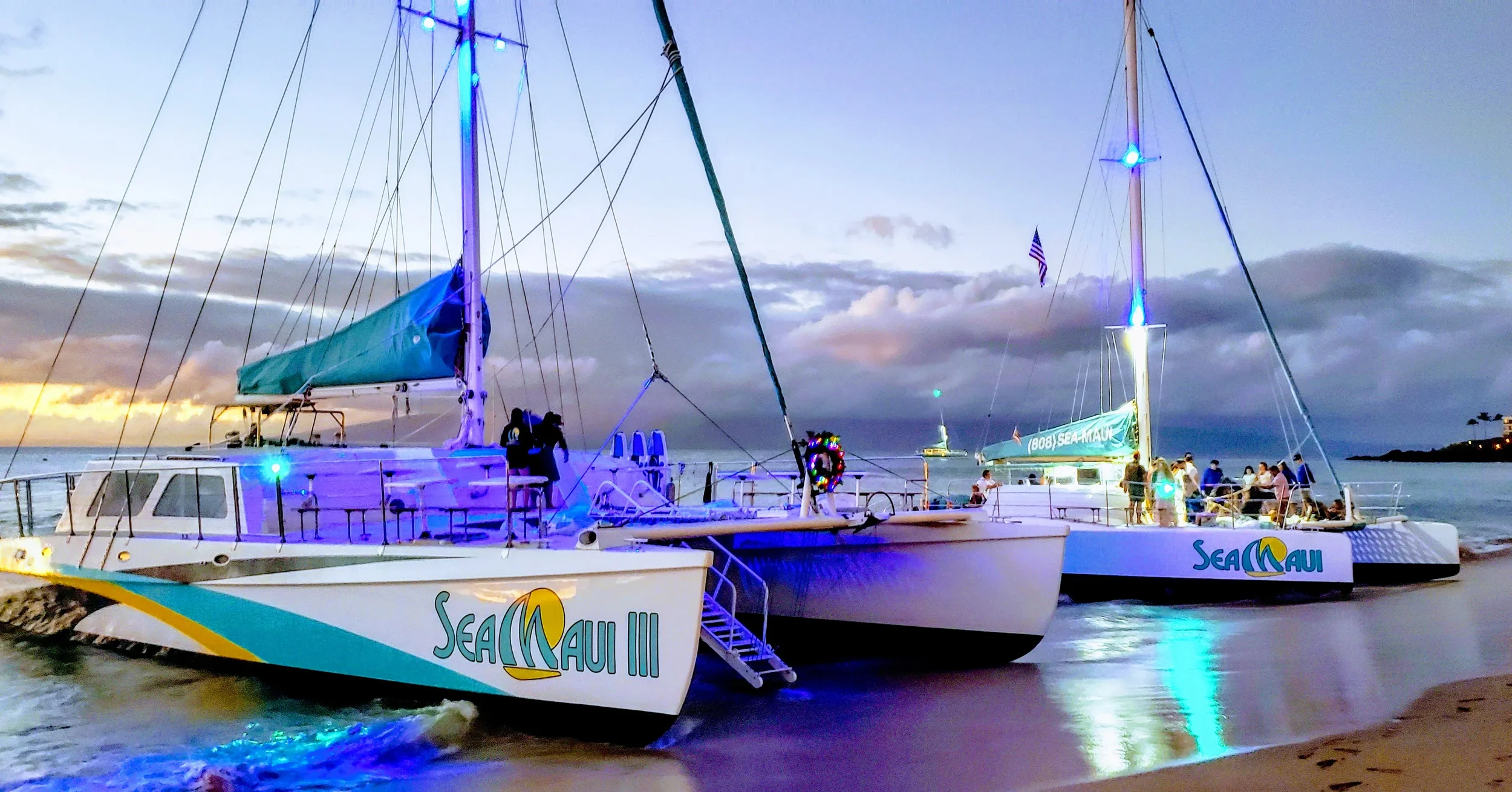 Two sailboats docked at a beach during sunset, with people on board and colorful lights illuminating the scene. The sailboats have the text 'Sea Maui' on their sides.