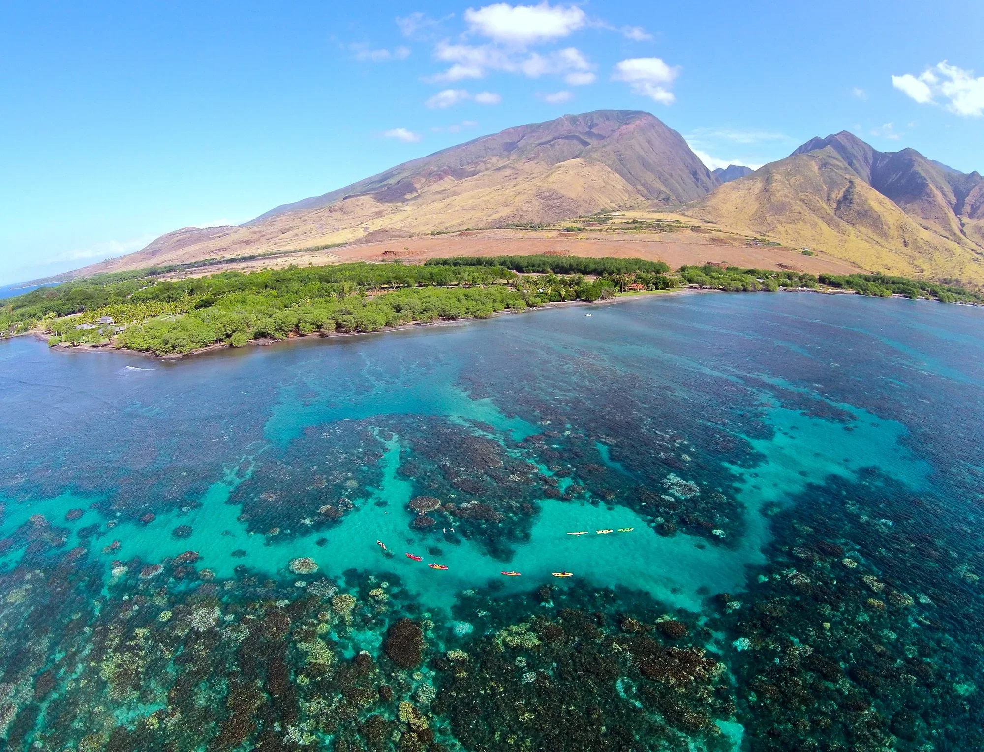 Aerial view of a tropical island with lush green forests, turquoise ocean waters, and a mountain range in the background under a partly cloudy sky.