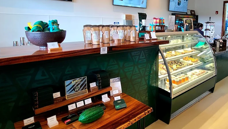 Display case with assorted pastries and desserts at a cafe counter, with bags of coffee and other items on the wooden counter behind it.