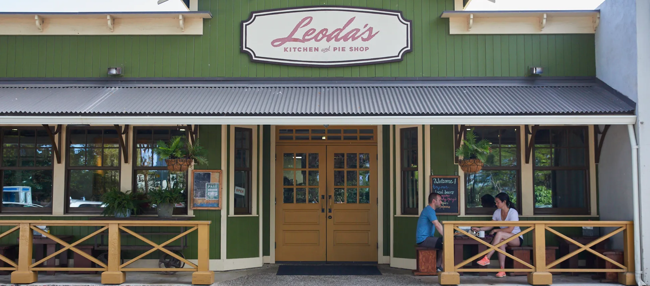 Exterior of Leah's Kitchen and Pie Shop, with two people sitting at a table on the front porch. The shop has a green facade, large windows, and a sign above the entrance. There are potted plants and hanging baskets outside.