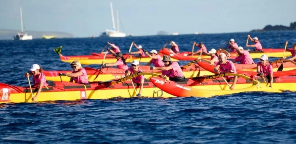 A group of people rowing yellow and red racing boats on a body of water, wearing pink shirts and white hats.
