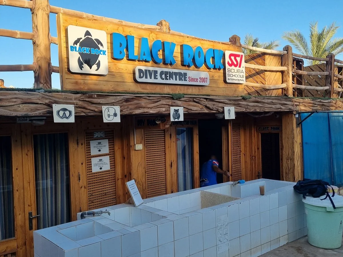 Wooden Emporium building with signs for Black Rock Dive Centre, SSI Scuba Schools International, and symbols for snorkeling, diving, and gear. Palm trees in background, blue skies, and a person wearing a blue shirt near the entrance.