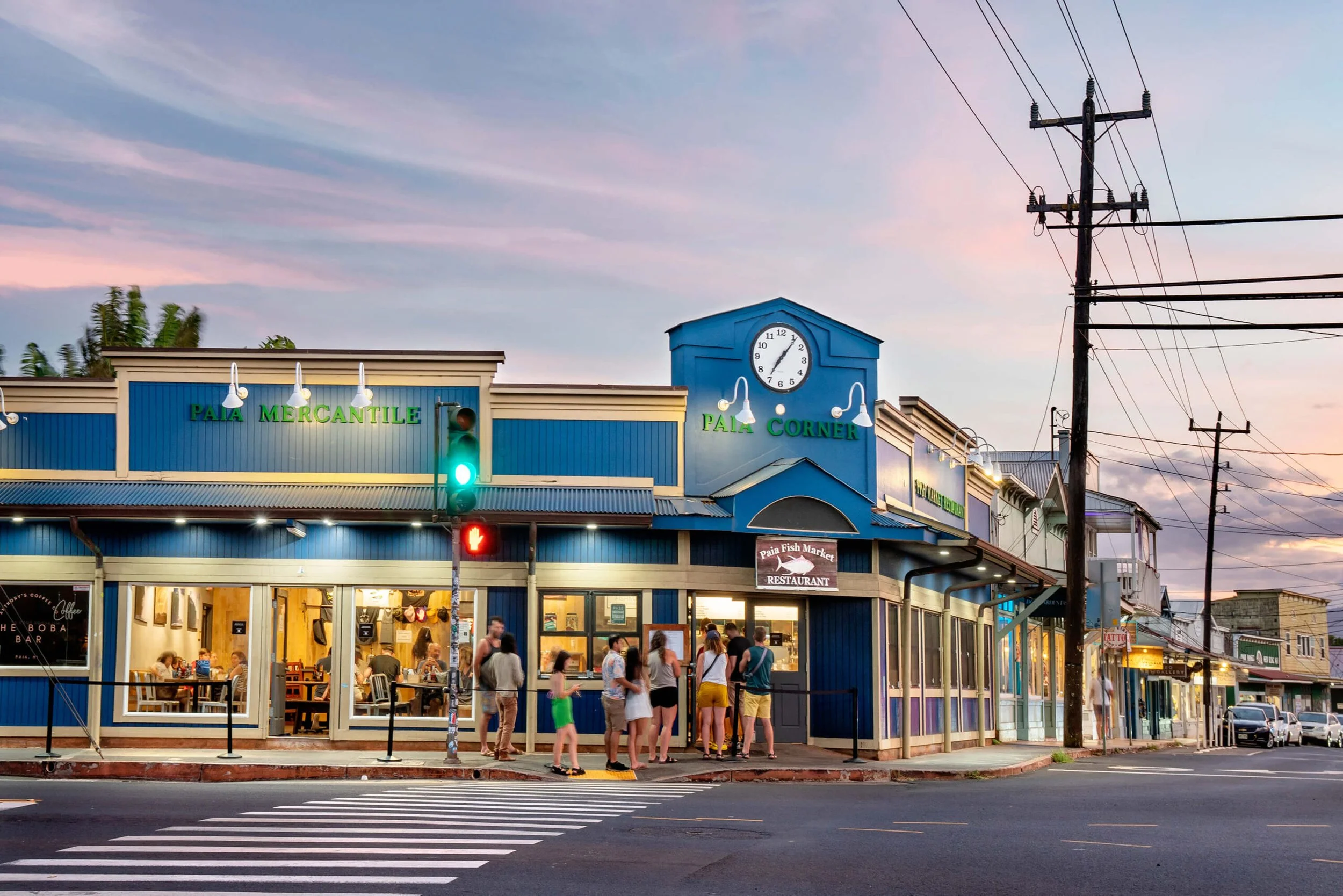 Street scene at sunset showing a blue restaurant building called 'Pala Corner' with a clock above the entrance, customers lining up outside, and traffic lights at the intersection.