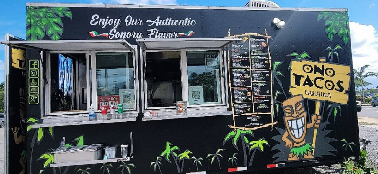 A food truck with a tropical theme featuring green leaves and a smiling cartoon Tiki figure holding a sign that says 'ONO TACOS LAHAINA.' The truck's slogan reads 'Enjoy Our Authentic Sonora Flavor,' with a menu board on the side. The window has a small counter with condiments and a water bottle.