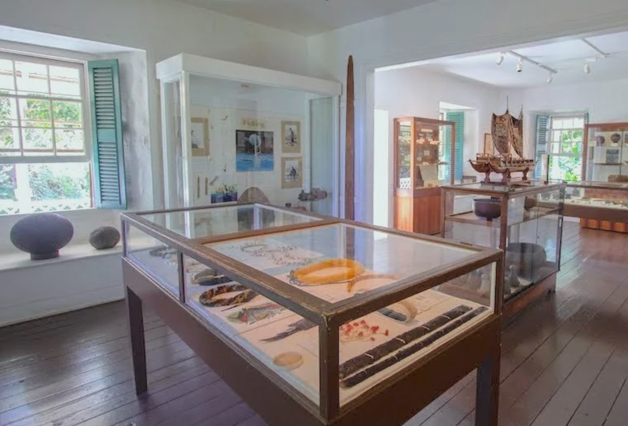 A museum exhibit featuring glass display cases with artifacts, pottery, and models of ships, within a room with white walls, wooden floors, and windows with open shutters.