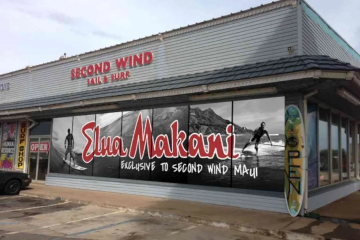 The storefront of a surf shop named Euma Makani with a large window display. The display features a promotional poster with images of surfers in black and white, a mountain in the background, and the shop's name written in large red letters. There is a surfboard sign leaning against the wall outside the shop and an open sign on the door.