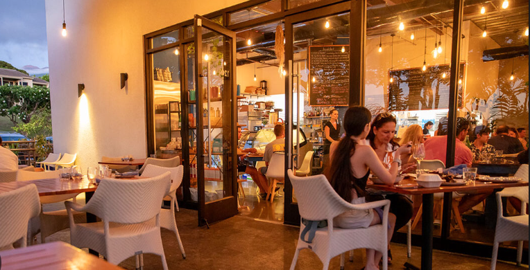 People dining inside a cozy restaurant with large glass windows and an outdoor patio area.