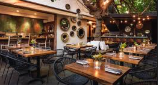 Outdoor patio of a restaurant with wooden tables set with plates, glasses, and silverware, surrounded by black chairs. String lights hang above, and there is a tree with green leaves in the background.