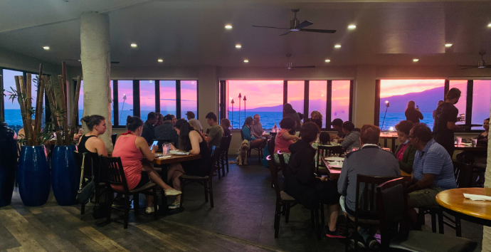 Interior of a restaurant filled with people dining and socializing during sunset, large windows showing purple and pink sky with mountains and palm trees in the background.