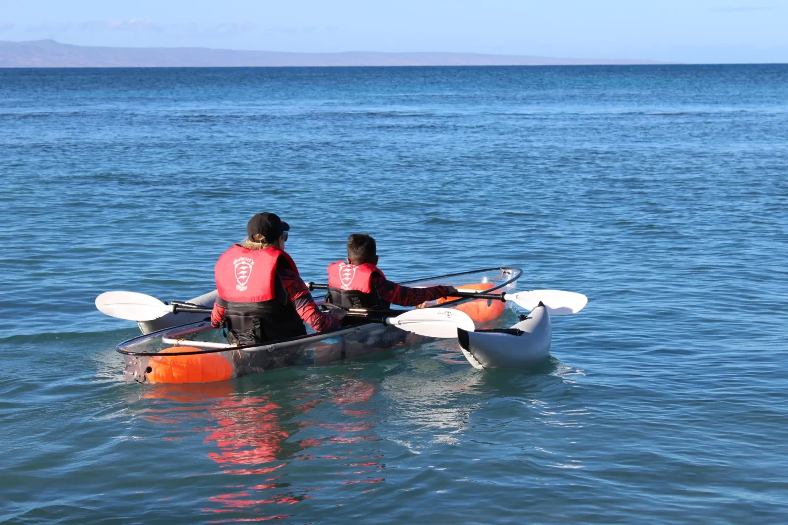 Two people paddling a tandem kayak on a calm body of water, wearing red life jackets. The scene is set in a sunny outdoor environment with a distant mountain range in the background.