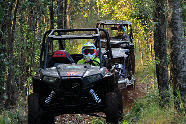 Two off-road utility vehicles driving through a dense forest trail. The person in the front vehicle is wearing a helmet and goggles, while the passenger in the back vehicle is also wearing a helmet and sunglasses.