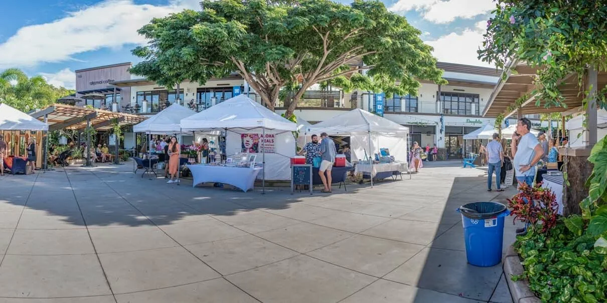 Outdoor marketplace with white tents, vendors, and shoppers in a plaza with trees and modern buildings in the background.