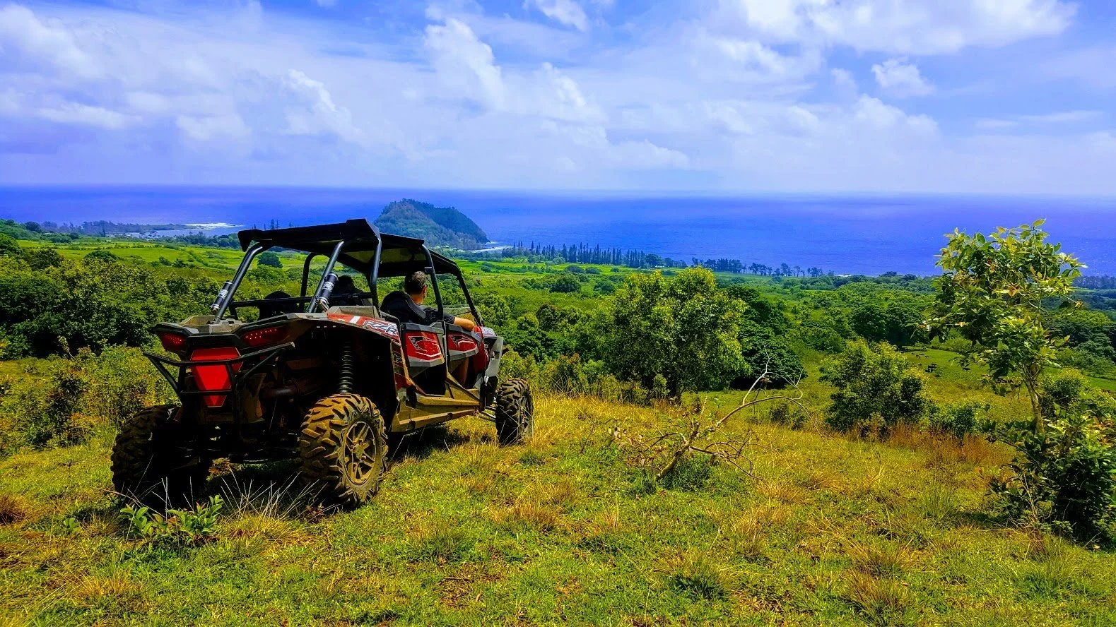 Off-road vehicle parked on a grassy hill overlooking lush green landscape and the ocean under a partly cloudy sky.