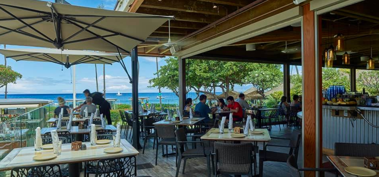 Outdoor restaurant with tables and chairs, some set with napkins and plates, overlooking the ocean with boats, trees, and a blue sky.