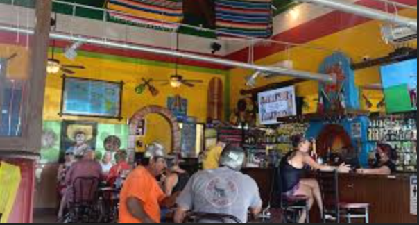 Inside a colorful restaurant with yellow walls, decorated with Mexican-themed items. Several people are sitting at tables and the bar, some wearing hats, with TVs mounted on the walls.