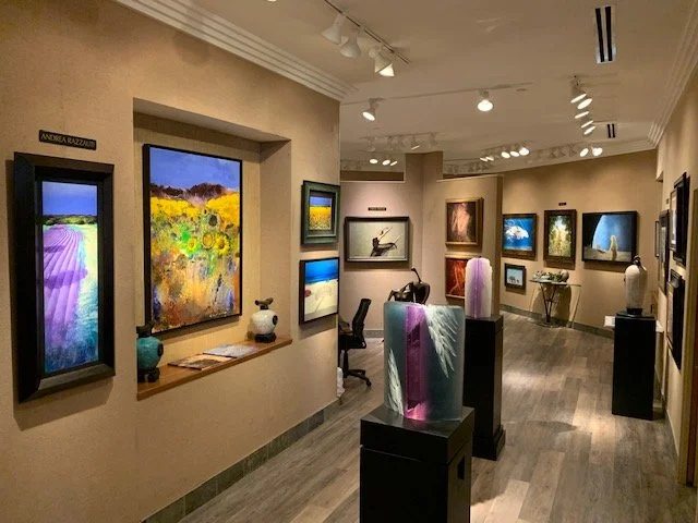 Art gallery with various paintings and sculptures on display, beige walls, wooden flooring, and track lighting.