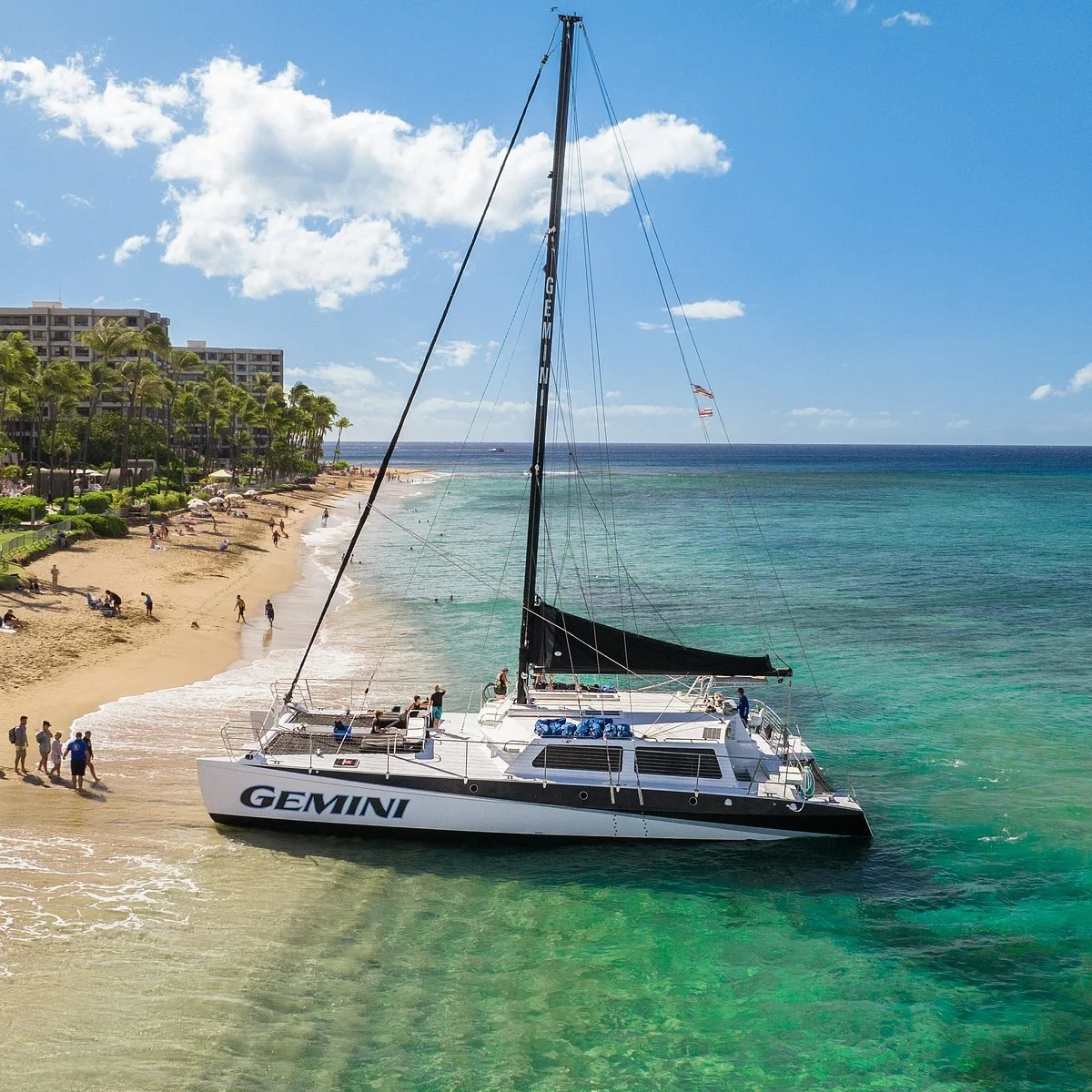 A white sailing yacht named Gemini with black sails is anchored near a sandy beach with people relaxing and walking. The background includes palm trees, a clear blue sky with some clouds, and buildings along the shoreline.