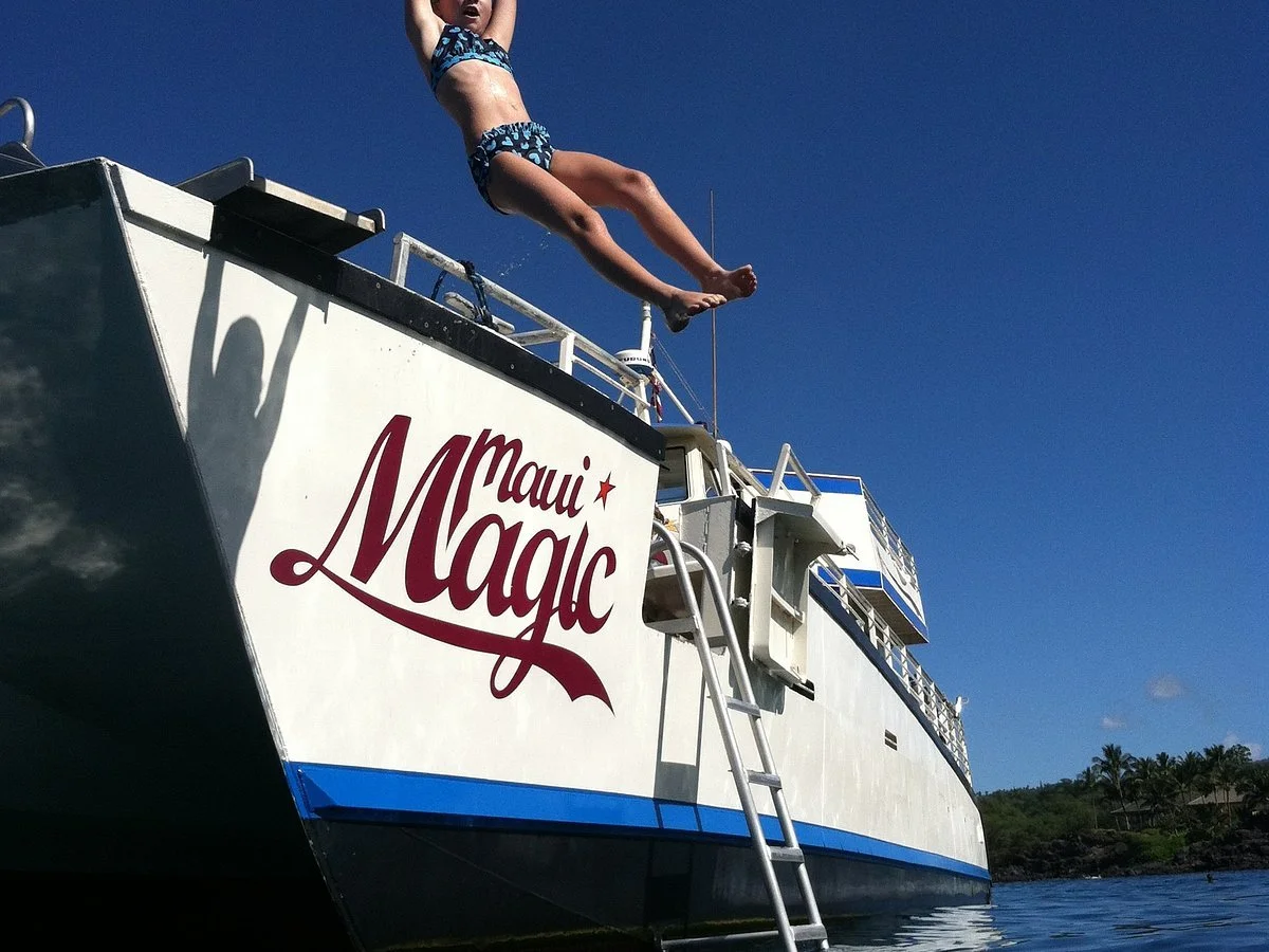 Child jumping off the side of a boat named Maui Magic into the water against a clear blue sky.