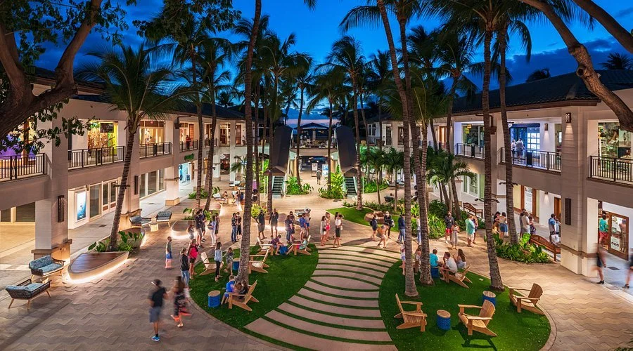 Outdoor courtyard of a tropical hotel with palm trees, balcony rooms on two levels, and people walking and socializing in the evening.
