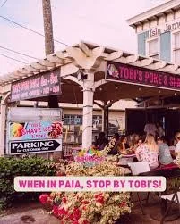 Outdoor scene of Tobi's Deli & Cafe with a pink sign, people sitting at tables, and a parking lot in front with floral decorations.