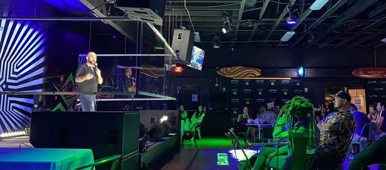 A stand-up comedian performs on stage at a nightclub with an audience seated. The stage has a microphone and lighting effects, with some screens and colorful decor.