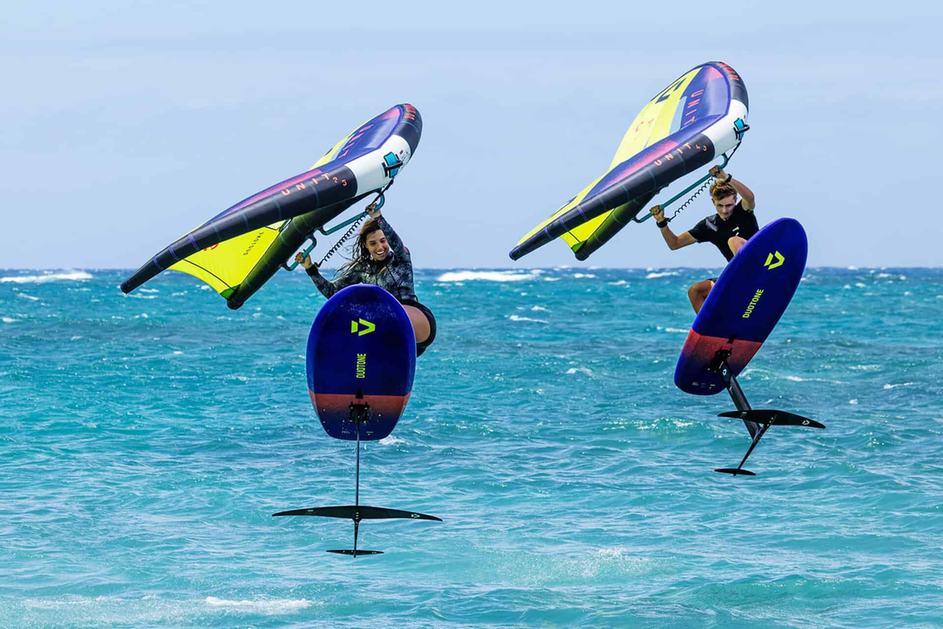 Two people on hydrofoil boards flying above the water, each holding a colorful sail, with a blue ocean and sky in the background.