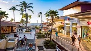 Outdoor shopping area with palm trees, stores, and people walking, under a blue sky