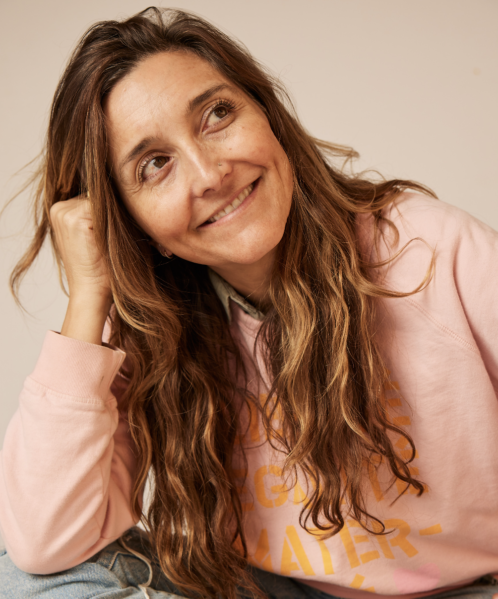 A woman with long, wavy brown hair wearing a pink sweatshirt and smiling, resting her head on her hand against a plain background.