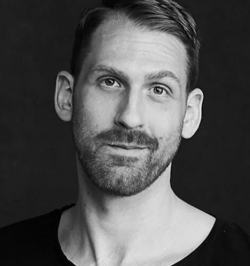 Black and white close-up portrait of a man with short hair, a beard, and a slight smile, against a dark background.
