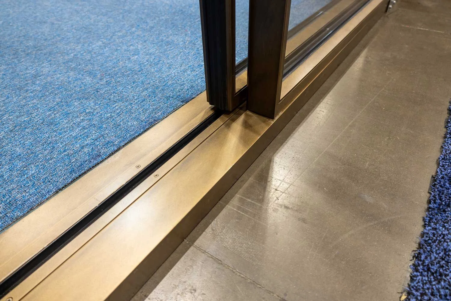 Close-up of a sliding glass door track on a brass threshold, with a blue carpet on the inside and a concrete floor outside.