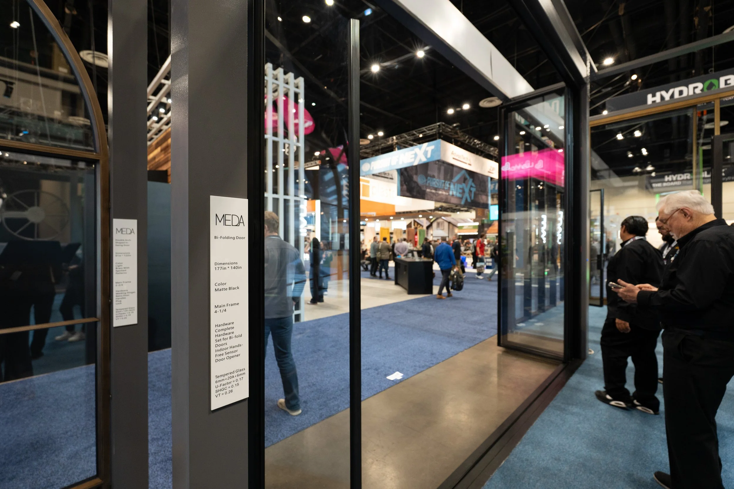 A trade show booth displaying a black bi-fold door labeled 'MEDA' with details about dimensions, color, and hardware. The booth is in a large exhibition hall with people walking and viewing various displays and booths.