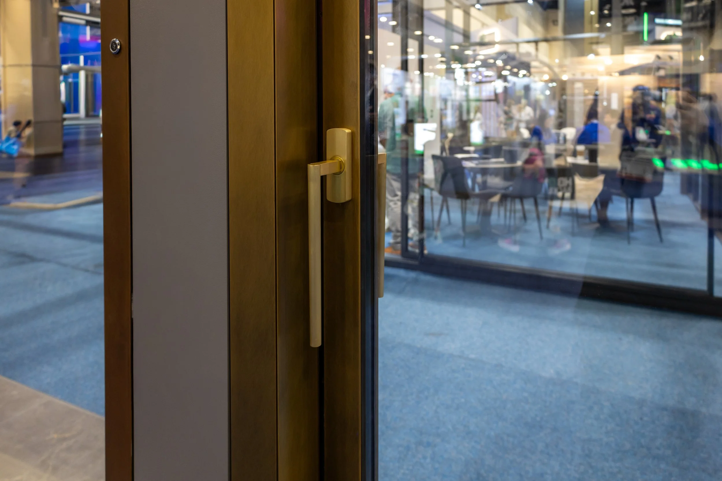 A glass door with a gold handle, showing an interior space with tables and chairs and a busy public area outside.