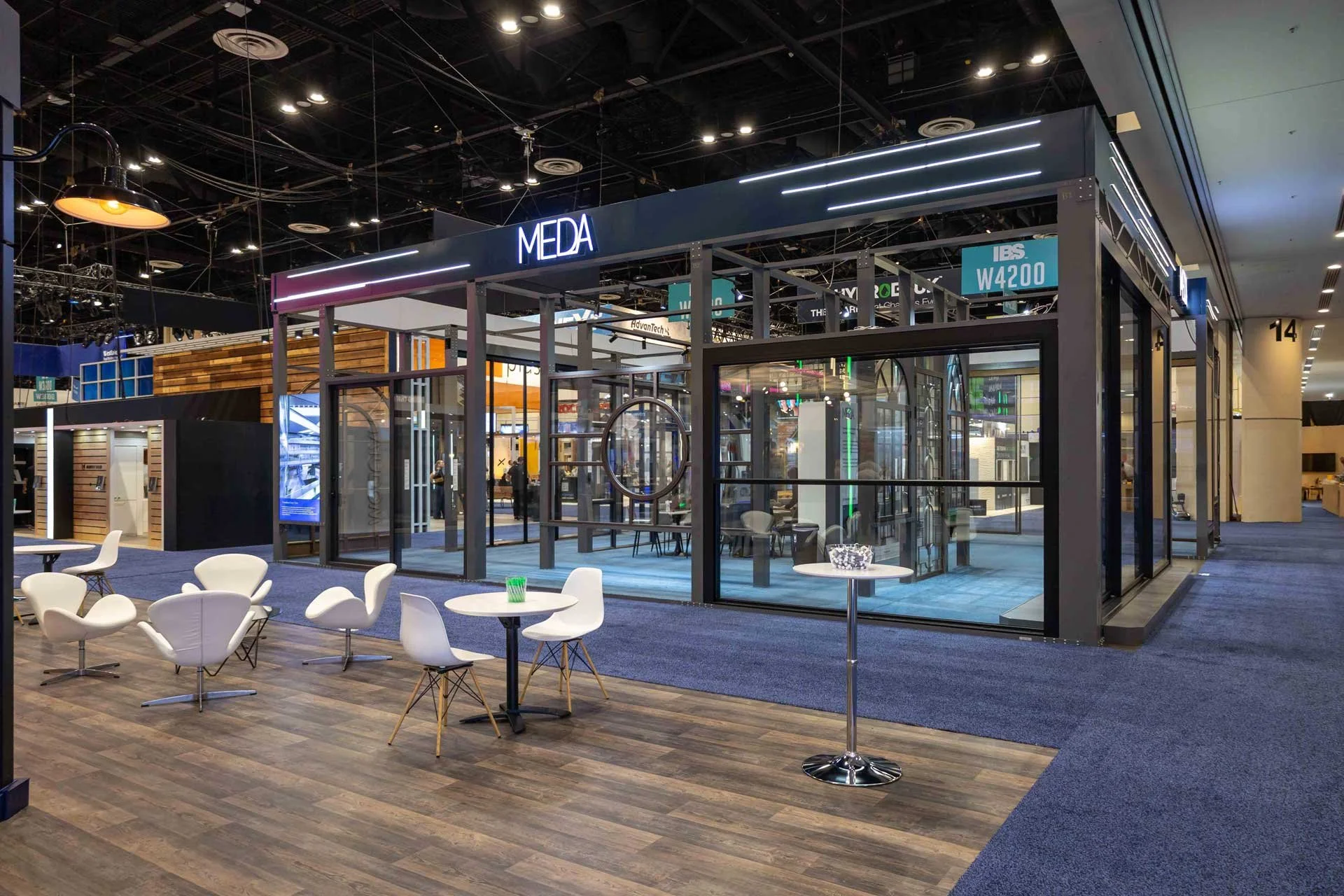 Trade show booth with the sign 'MEDA', glass walls, white chairs, and tables inside and outside on wooden and blue carpeted flooring.