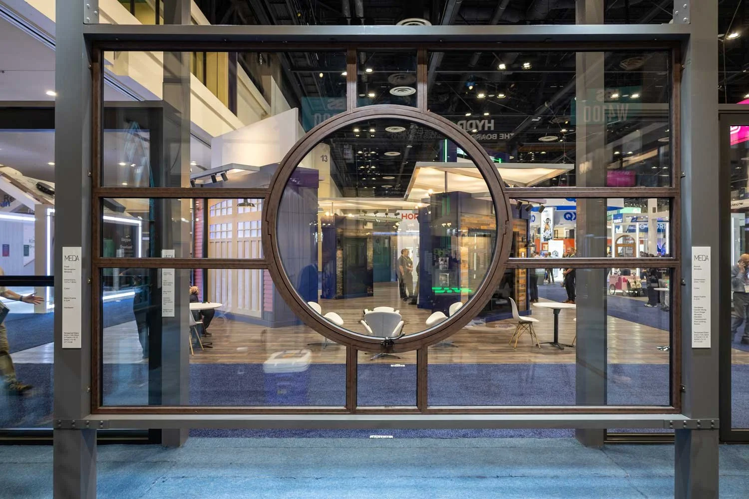 View through a large window with a circular design in the center, showing a modern interior space with seating and people inside a showroom or exhibition hall.
