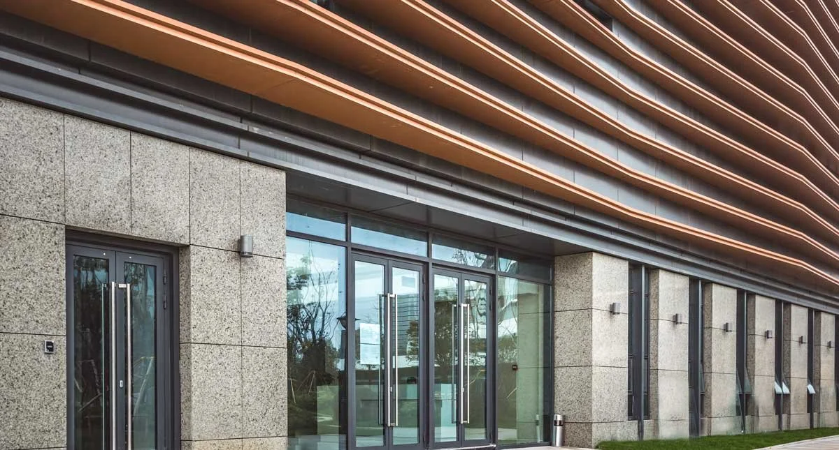 Modern commercial building with glass doors, granite walls, and horizontal metal and wooden paneling, with outdoor lighting fixtures.