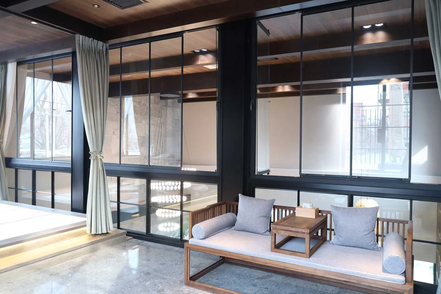 Interior view of a modern room featuring a window with beige curtains, a wooden bench with cushions, and glass walls with black metal framing.