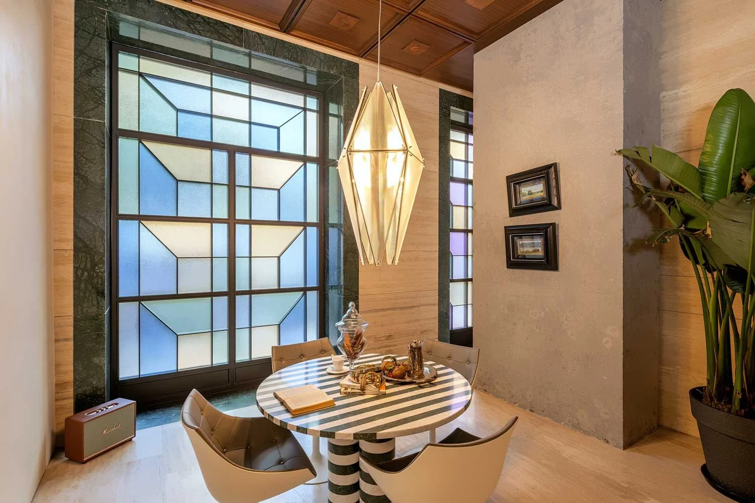 A cozy dining area with a round striped black and white table, surrounded by four modern beige chairs. A large, geometric pendant light hangs above the table. The background features a colorful stained glass window with blue, green, and purple sectio