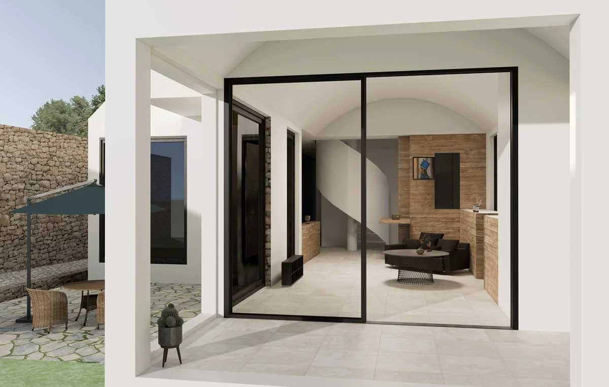 Modern interior view through sliding glass doors showing a living space with wooden features, a black sofa, and a small table. Part of an outdoor patio with chairs and an umbrella is visible to the left.