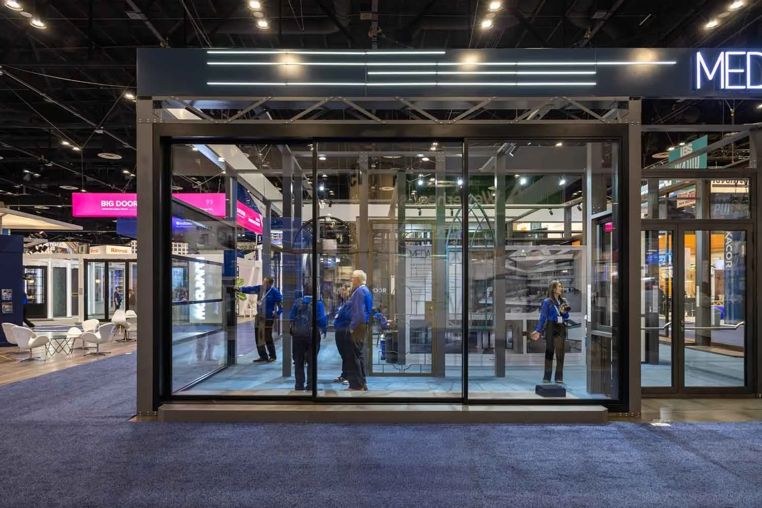 A glass-enclosed display booth at a trade show or convention, with people inside and outside. The booth has black framing and indoor lighting, with people engaged in conversation and a woman standing on a platform or step at the right side.
