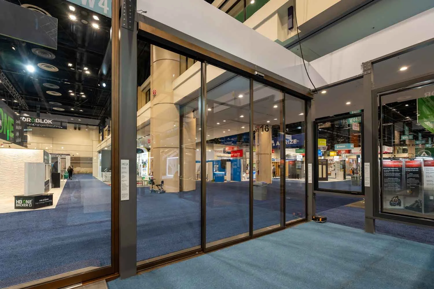 Glass door entrance at a convention center or exhibition hall with a view of an empty corridor and various booths in the background.
